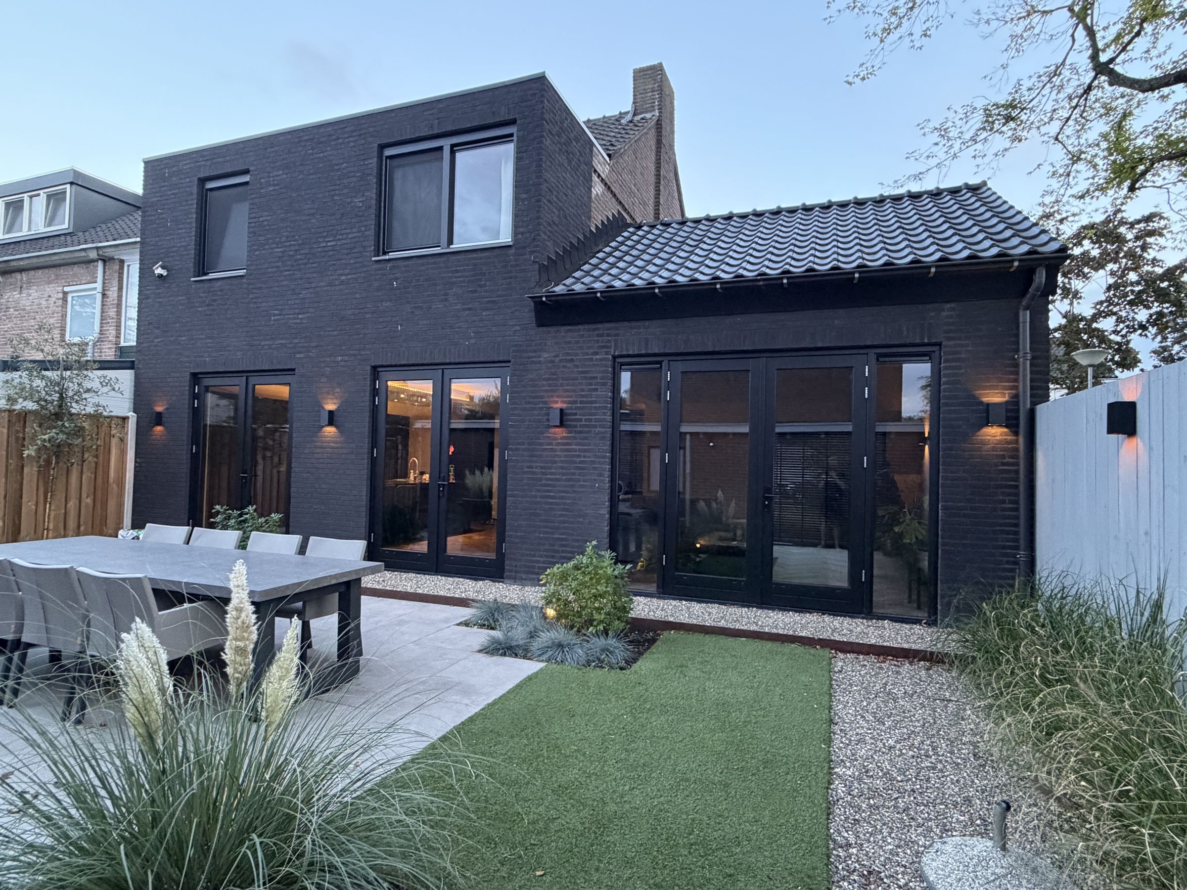 Opgeleverde woning met zwarte baksteen, zwarte kozijnen en een aanbouw, gezien vanuit de tuin in de avond