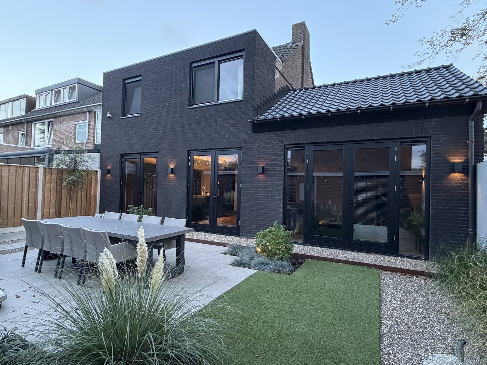 Achtergevel van een woning met zwarte baksteen, zwarte kozijnen en een aanbouw, gezien vanuit de tuin